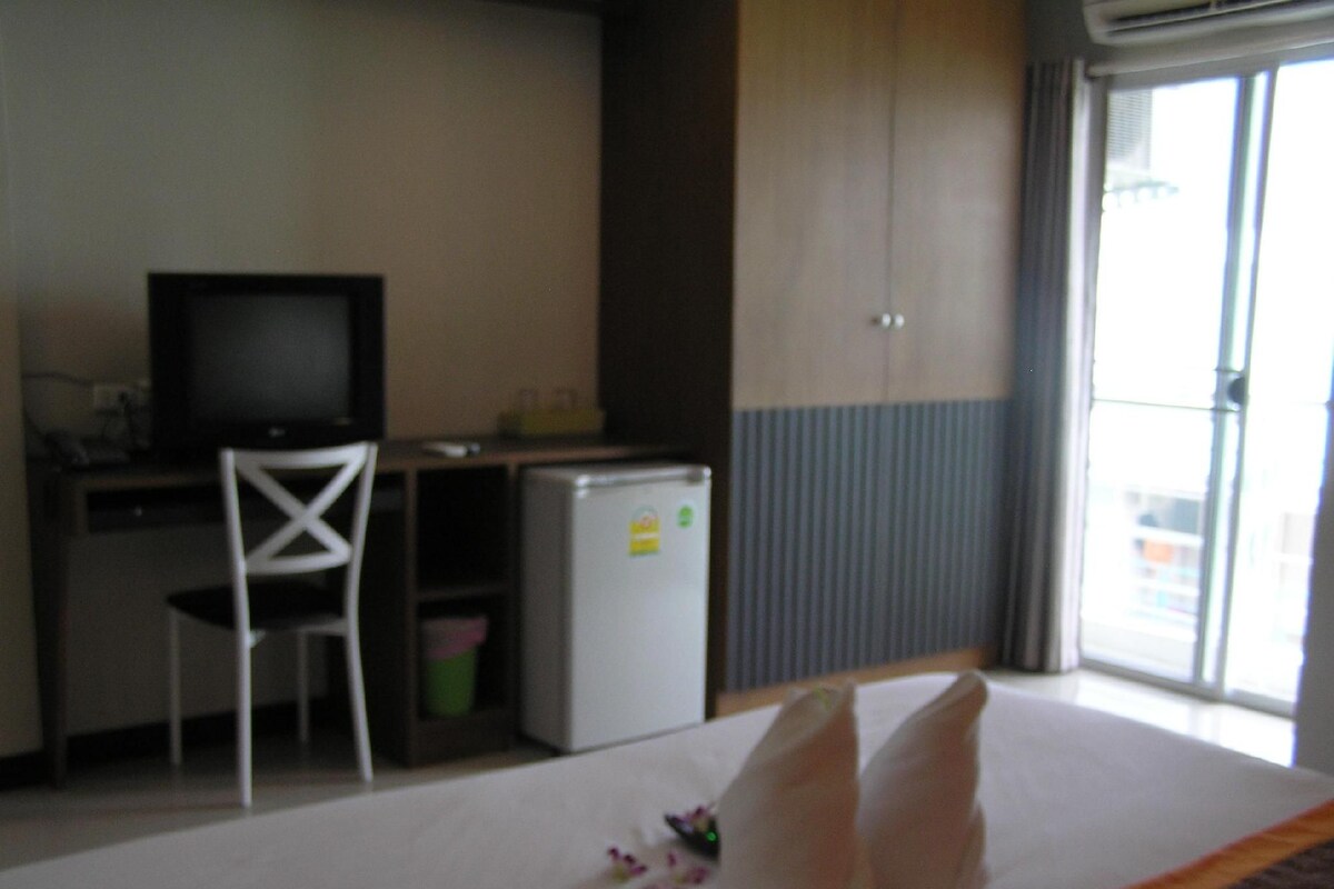 A hotel room with a bed, a desk with a computer, a chair, a small refrigerator, and a sliding door to the balcony.