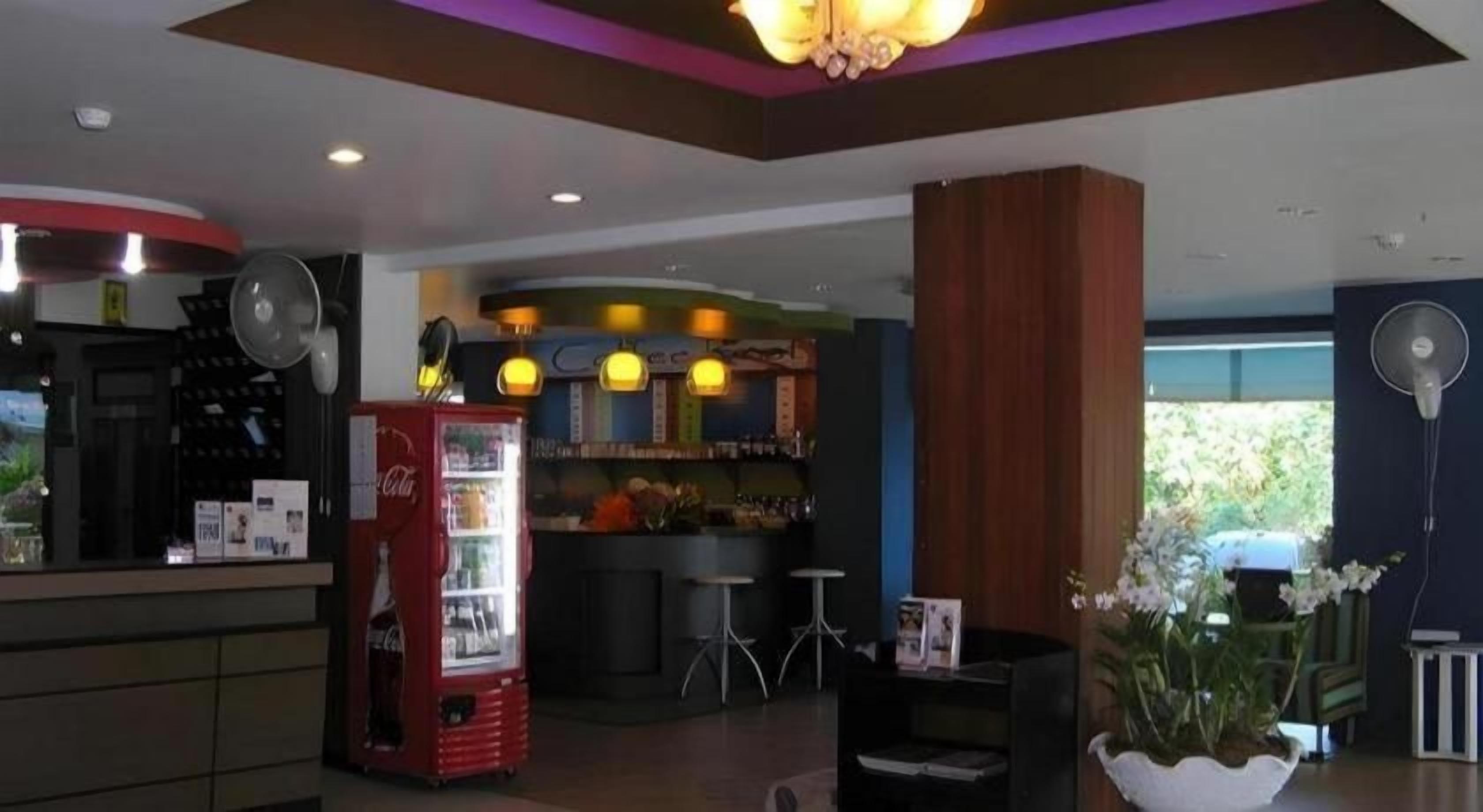 A hotel lobby with a reception desk, a Coca-Cola refrigerator, a bar area, and a seating area with a fan.