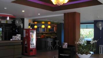 A hotel lobby with a reception desk, a Coca-Cola refrigerator, a bar area, and a seating area with a fan.
