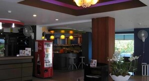 Un vestíbulo de hotel con mostrador de recepción, refrigerador de Coca-Cola, zona de bar y área de descanso con ventilador.