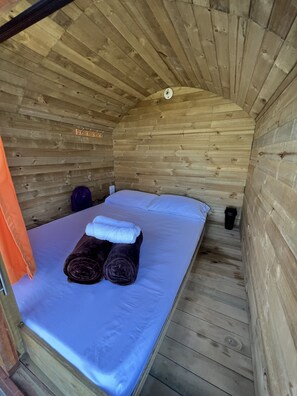 A small wooden cabin room with a bed, two rolled towels, a purple bag, and a black cup on the floor. - Loma Nevada Minca Hotel (Santa Marta)
