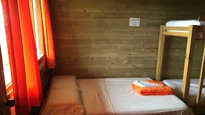 Una cama individual con colchón blanco y una manta naranja doblada, literas de madera a la derecha y una ventana con cortinas rojas.