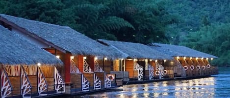 A row of overwater bungalows with thatched roofs, connected by a walkway, set against a backdrop of dense forest and a calm body of water.