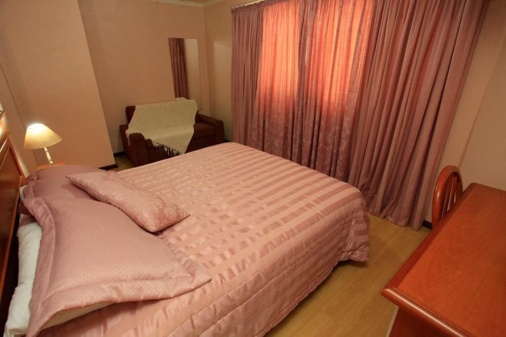 Habitación de hotel con cama, mesita de noche de madera, una silla y ventana con cortinas.