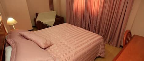 Habitación de hotel con cama, mesita de noche de madera, una silla y ventana con cortinas.