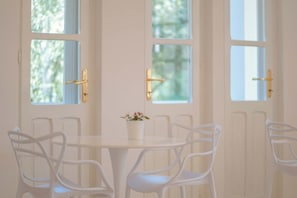 A room with a round table and four white chairs, a potted plant on the table, and three windows with wooden frames and gold handles. - Maison Del Sole (Quartu Sant'Elena)