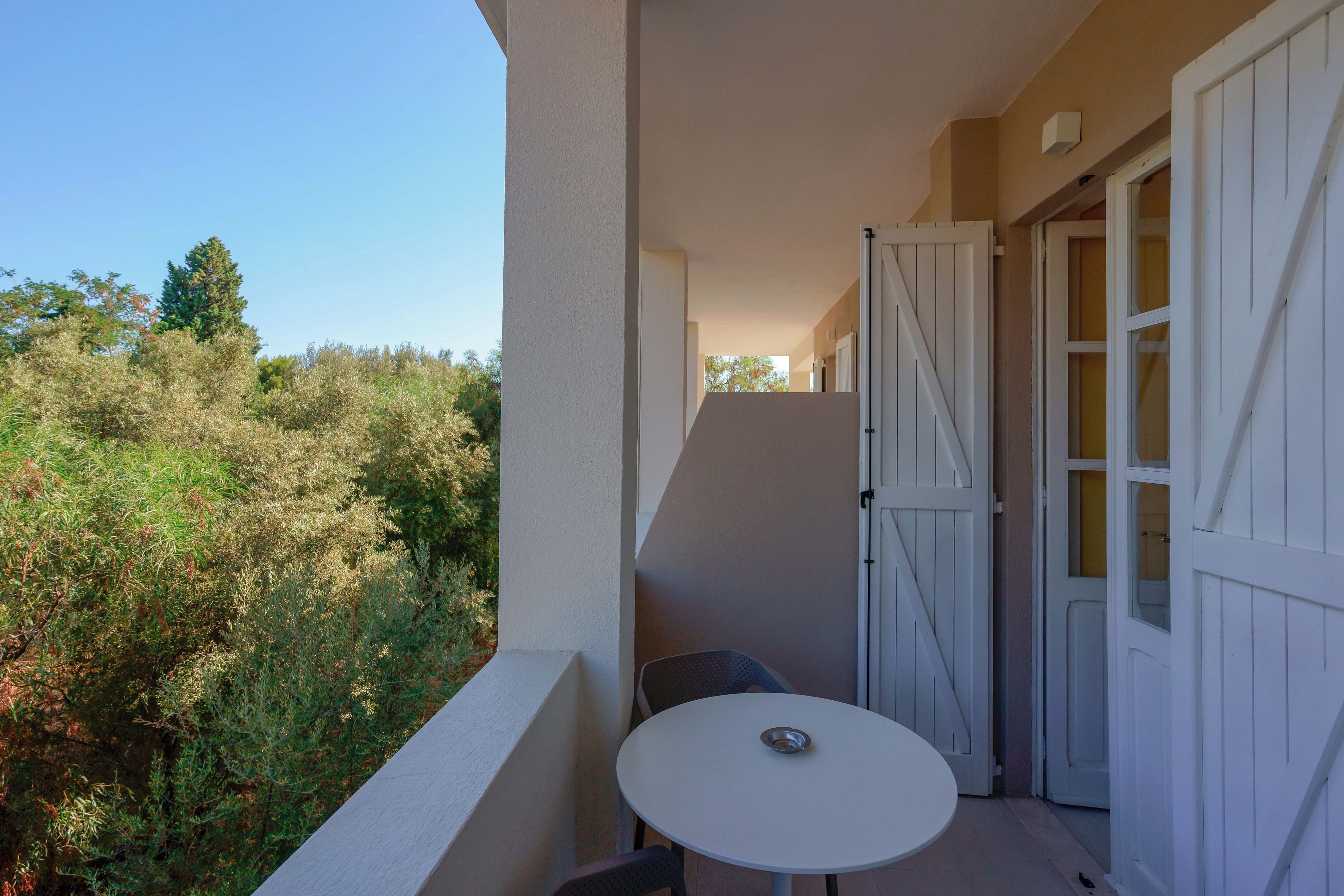 Un balcon avec une table et des chaises blanches, une porte blanche et une vue sur des arbres et des arbustes.