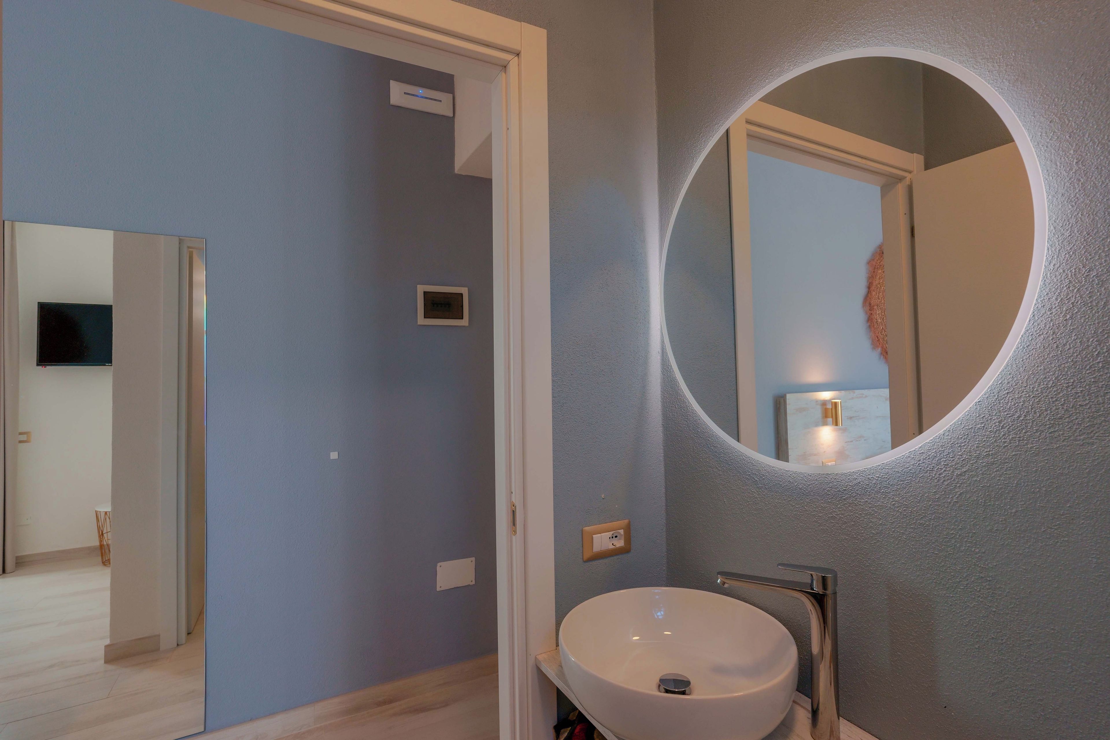 Une salle de bain avec un miroir rond, un lavabo et une porte donnant sur une autre pièce.