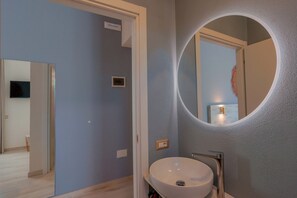 Un baño con un espejo redondo, un lavamanos y una puerta que da a otra habitación.