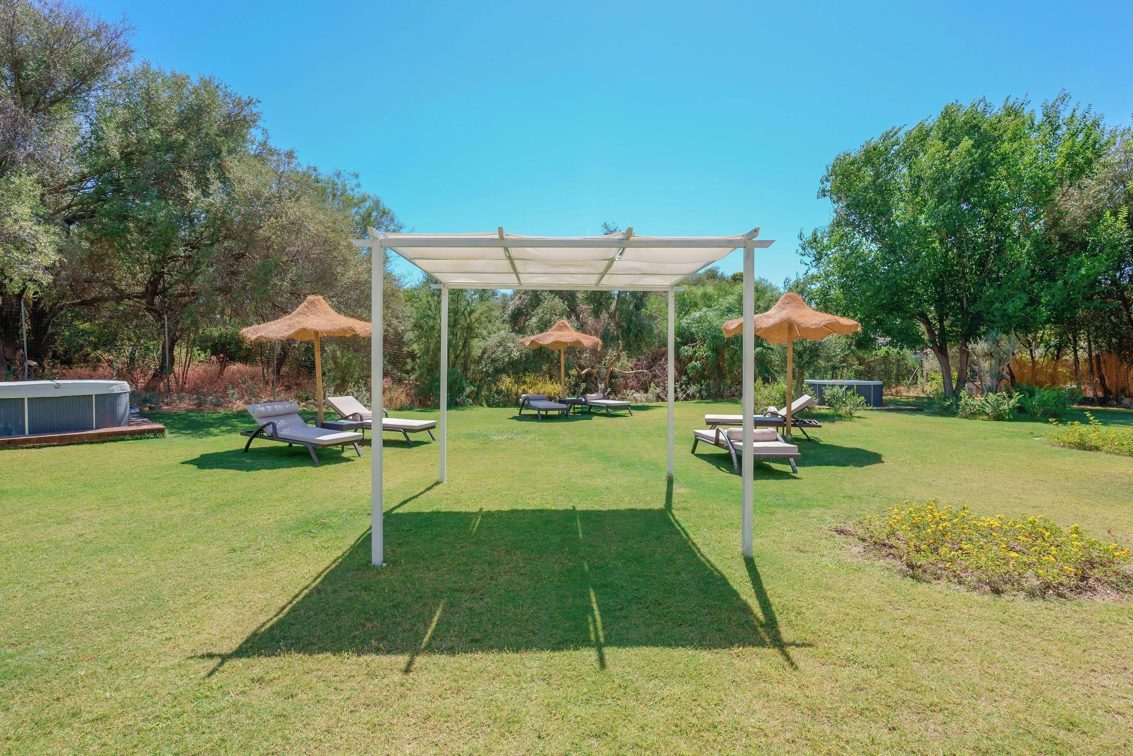 A covered outdoor seating area with lounge chairs and umbrellas, surrounded by trees and a hot tub.