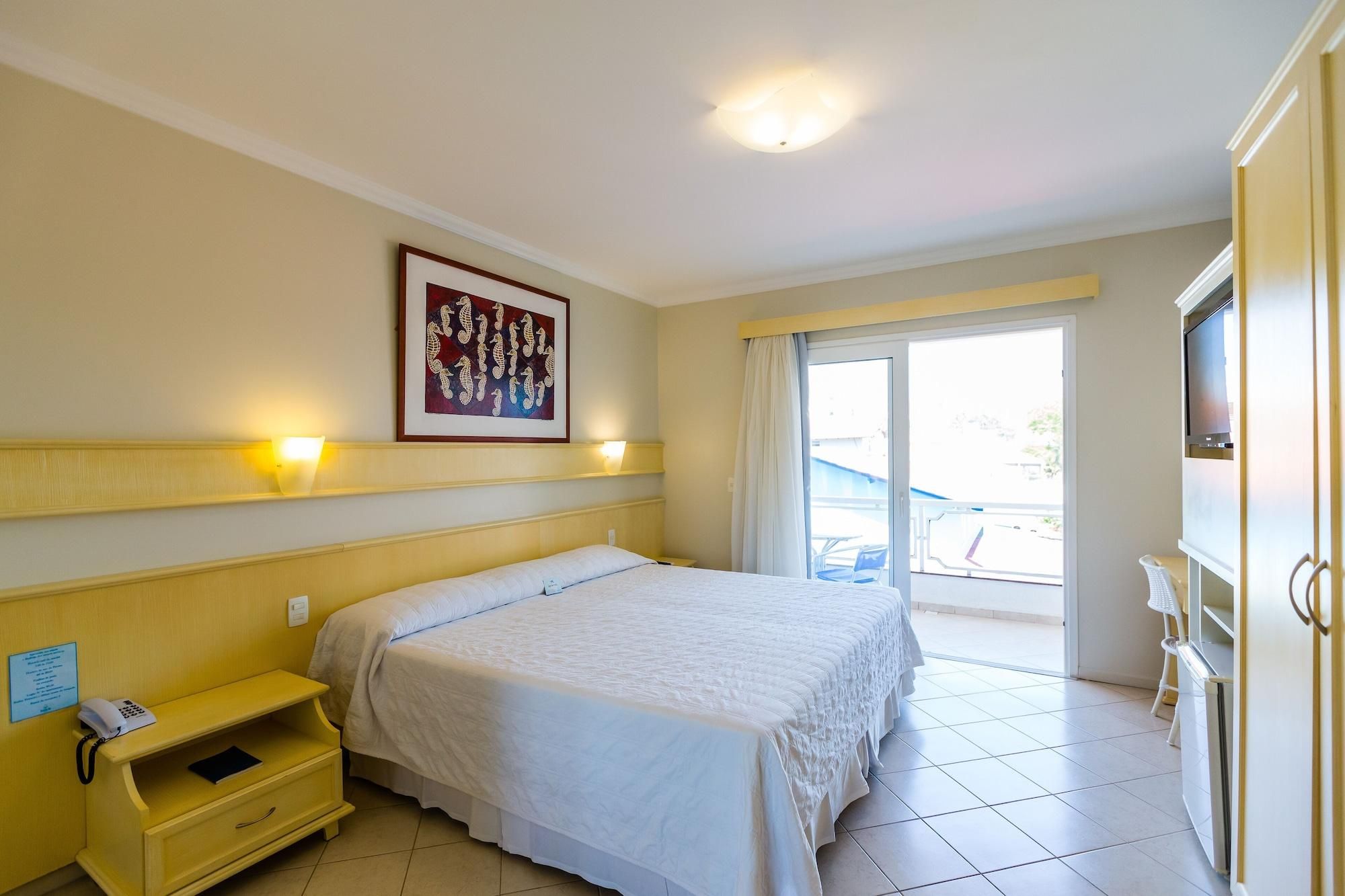 A hotel room with a bed, bedside table, lamp, artwork, and a view of the outside.