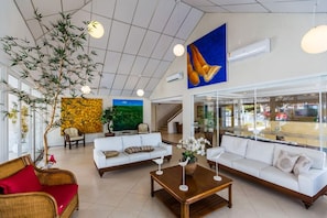 A spacious lobby with a large indoor plant, two white sofas, a wooden coffee table, and a large painting on the wall.