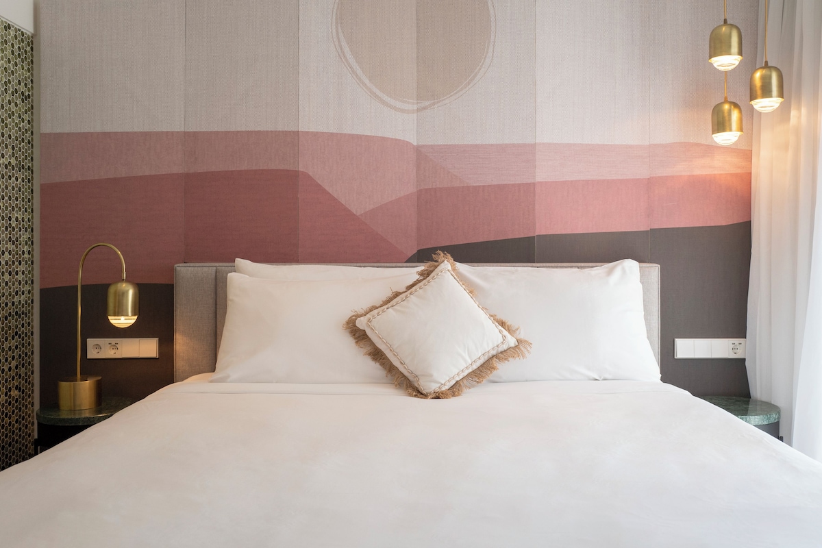 Een hotelkamer met een bed, een nachtkastje met een lamp en een muur met een geometrisch patroon.