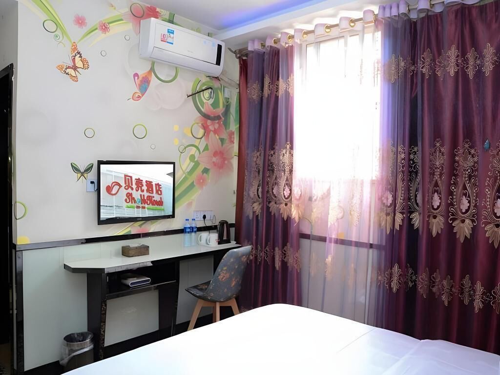 A hotel room with a bed, a desk with a chair, an air conditioner, a TV, and decorative curtains.