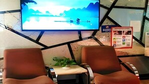 A lobby with a large TV screen displaying a scenic view, two brown chairs, a small table with a plant, and a poster on the wall. - Shell Shanghai Jiading District Tongji University Xinhuang Road Hotel (Shanghai)