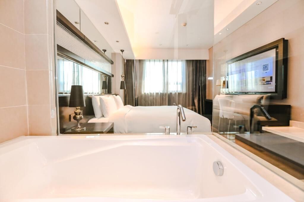 A modern hotel room with a large bathtub, a bed, a bedside table with a lamp, and a TV mounted on the wall.