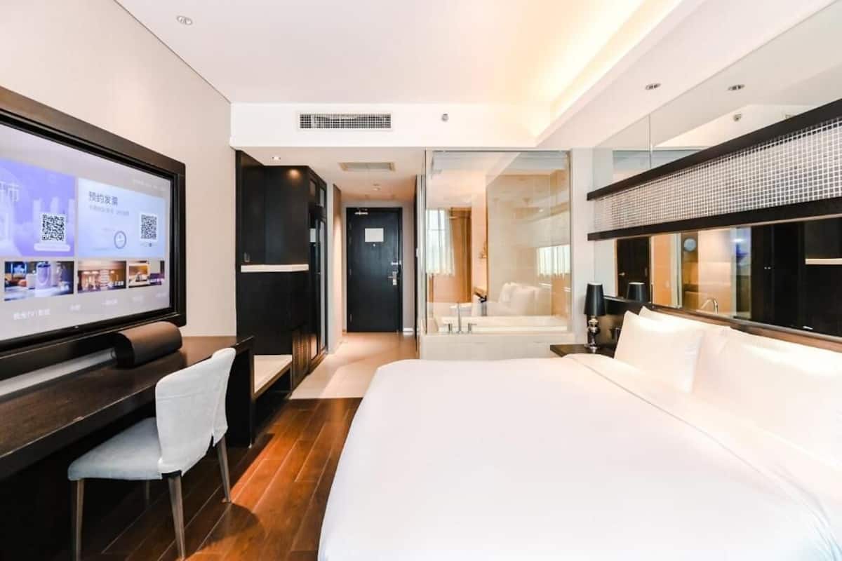 A modern hotel room with a large bed, a desk with a computer, a TV, and a glass-enclosed bathroom.