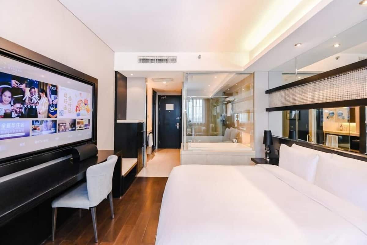 A modern hotel room with a large bed, a TV, a desk, and a glass partition leading to a bathroom.