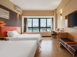 Een hotelkamer met twee bedden, een bureau, een stoel, een televisie en een raam met uitzicht op de stad.
