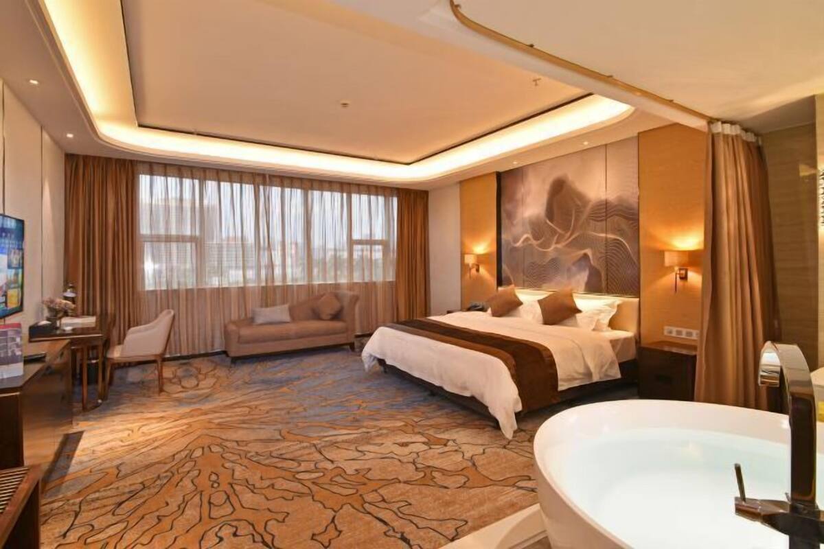 Ein Hotelzimmer mit einem grossen Bett, einer Sitzecke mit einer Couch, einem Schreibtisch mit einem Stuhl, einem grossen Fenster mit Vorhängen und einer Badewanne.