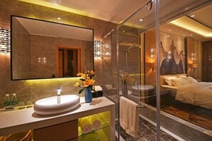 Une salle de bain moderne avec une douche en verre, un grand miroir et un lit visible à travers le verre.