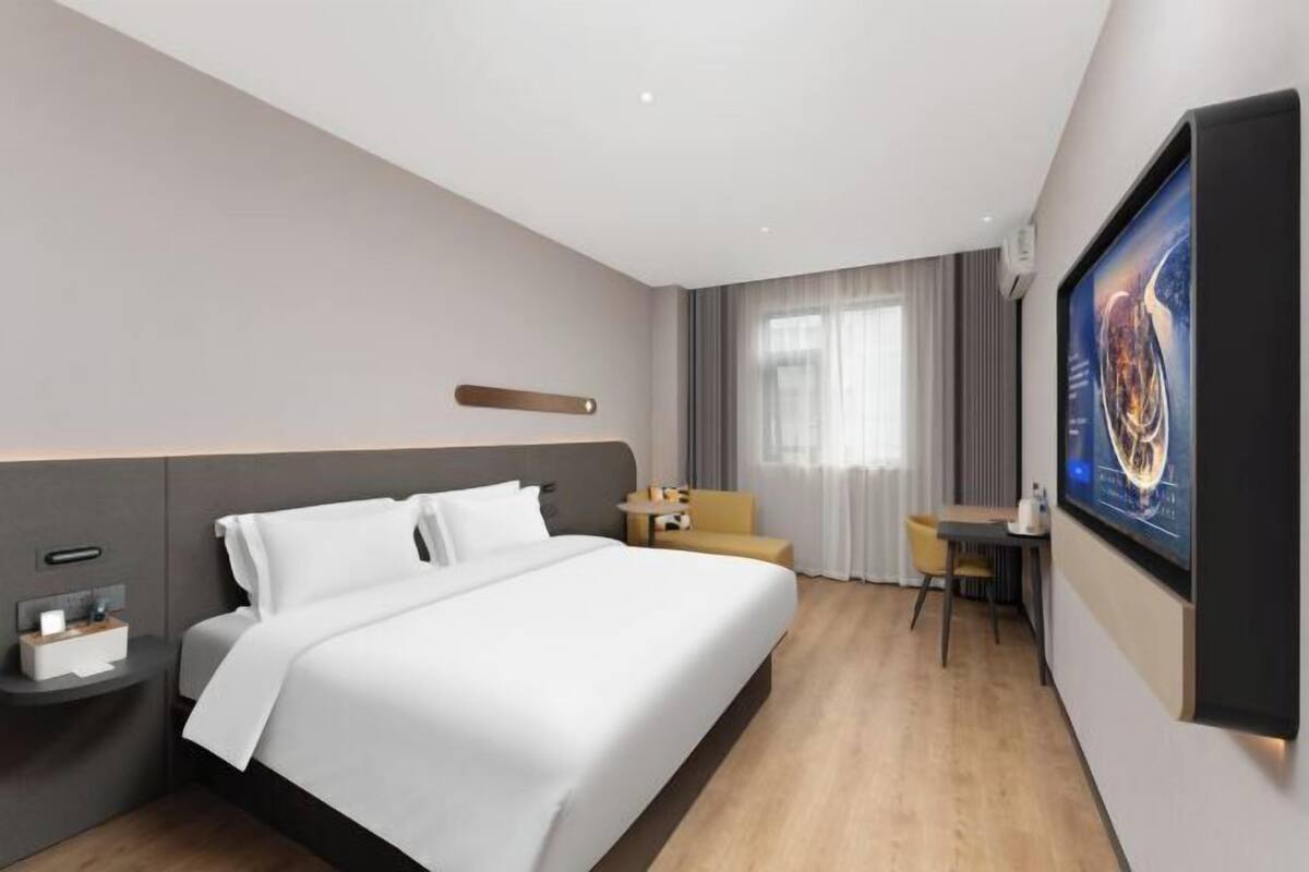 A hotel room with a large bed, a bedside table, a small desk, a chair, and a large TV screen.