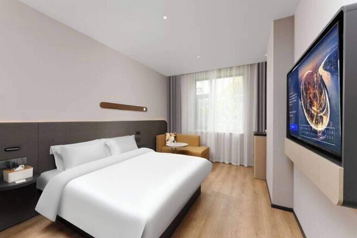 A modern hotel room with a large bed, a TV on the wall, a small table, and a window with curtains.