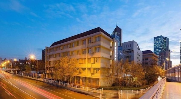 Un edificio de varios pisos con un diseño arquitectónico moderno, rodeado de árboles y una calle concurrida con vehículos en movimiento.