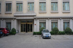 Un edificio con entrada de doble puerta, dos coches estacionados adelante y una pequeña señal en el suelo.