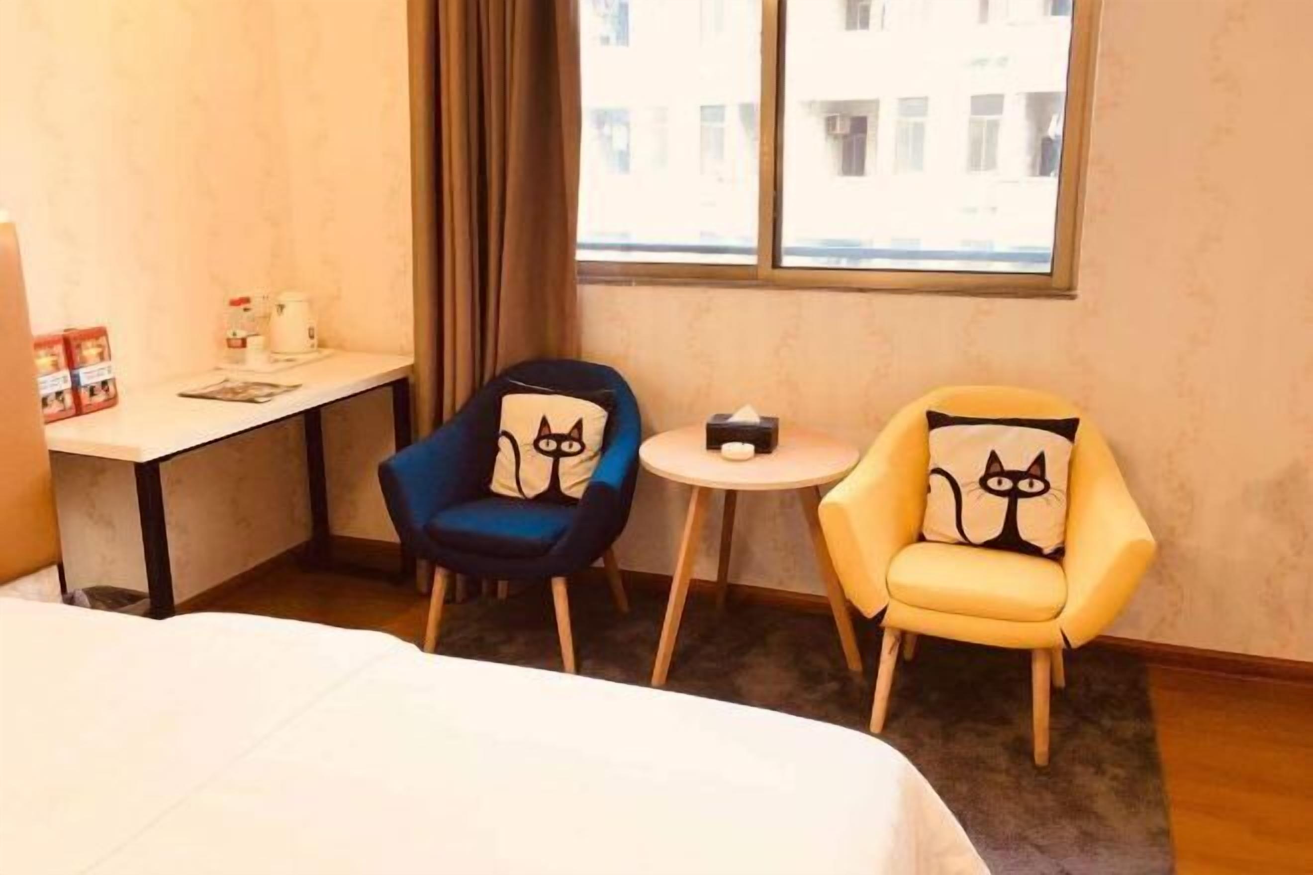 Habitación de hotel con cama, escritorio con silla, dos sillones con cojines con motivos de gatos, una mesa redonda pequeña y una ventana con vistas a edificios.