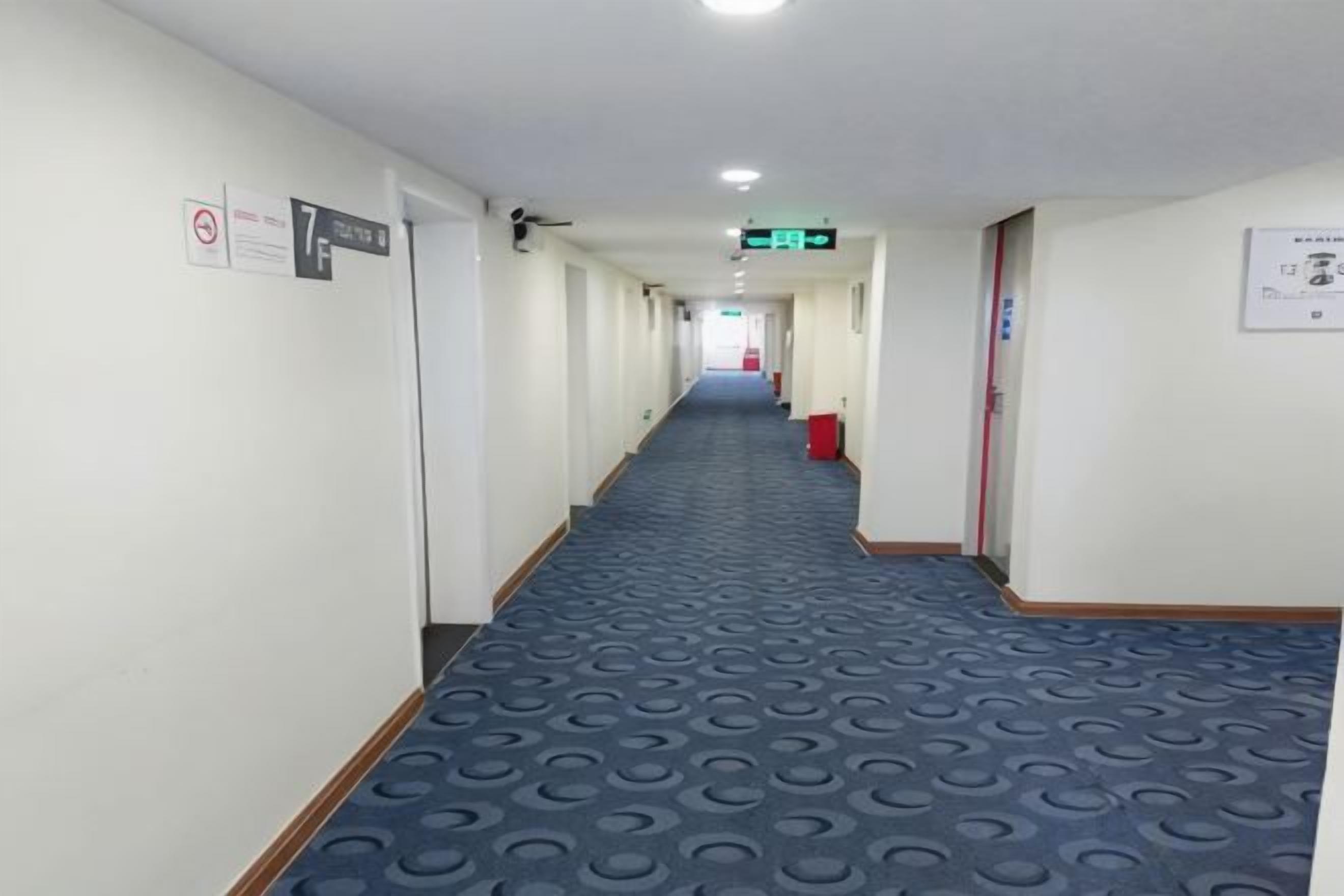 A hotel corridor with a blue patterned carpet, doors on the left, and a red trash bin on the right.
