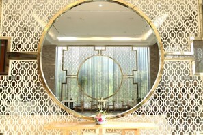 Une pièce dotée d’un grand miroir rond posé sur un piédestal en bois, d’un mur décoratif orné d’un motif géométrique et offrant une vue par la fenêtre.