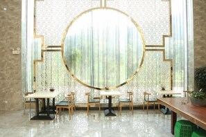 Un espace repas agrémenté d’un miroir rond, de tables, de chaises et offrant une vue à travers une fenêtre à motifs.