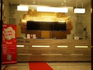 A hotel reception area with a modern design, featuring a large flat-screen TV, a red carpet, and a promotional sign.