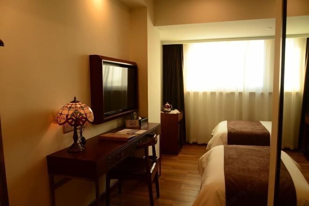 A hotel room with a bed, a desk with a lamp, a television, and a window with curtains.