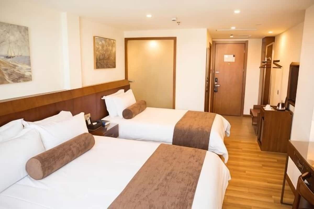 Une chambre d’hôtel avec deux lits, une tête de lit en bois, un bureau, une chaise, un téléviseur et une porte donnant sur une autre pièce.