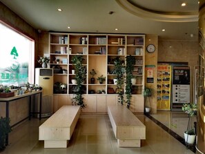 Un espace d’accueil comprenant un comptoir de réception en bois, deux bancs en bois, une bibliothèque remplie de livres et de plantes, une horloge murale et une affiche avec du texte.