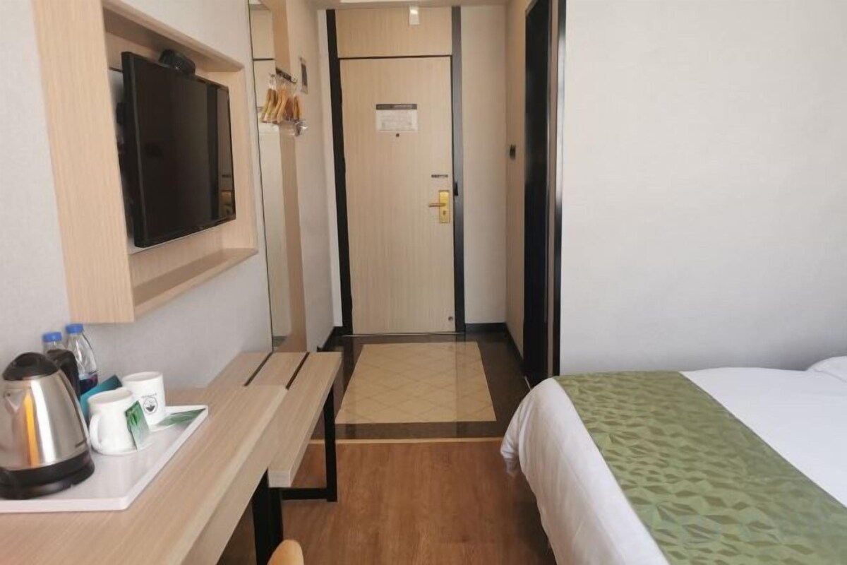 Ein Hotelzimmer mit einem Bett, einem Schreibtisch mit Wasserkocher und Tassen, einem Fernseher und einer Tür zu einem Flur.