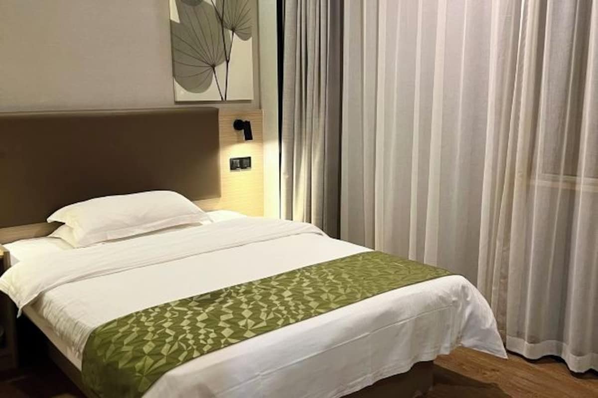Un lit bien fait, recouvert d’un plaid à motifs géométriques verts, un interrupteur fixé au mur et de fines rideaux transparents.