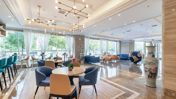 A spacious hotel lobby with a central round table, comfortable seating, and large windows offering a view of the outdoors.