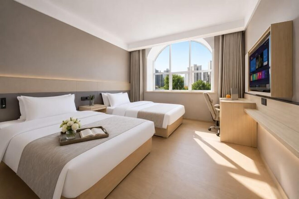 Habitación de hotel con dos camas, un escritorio, un televisor y un ventanal grande.