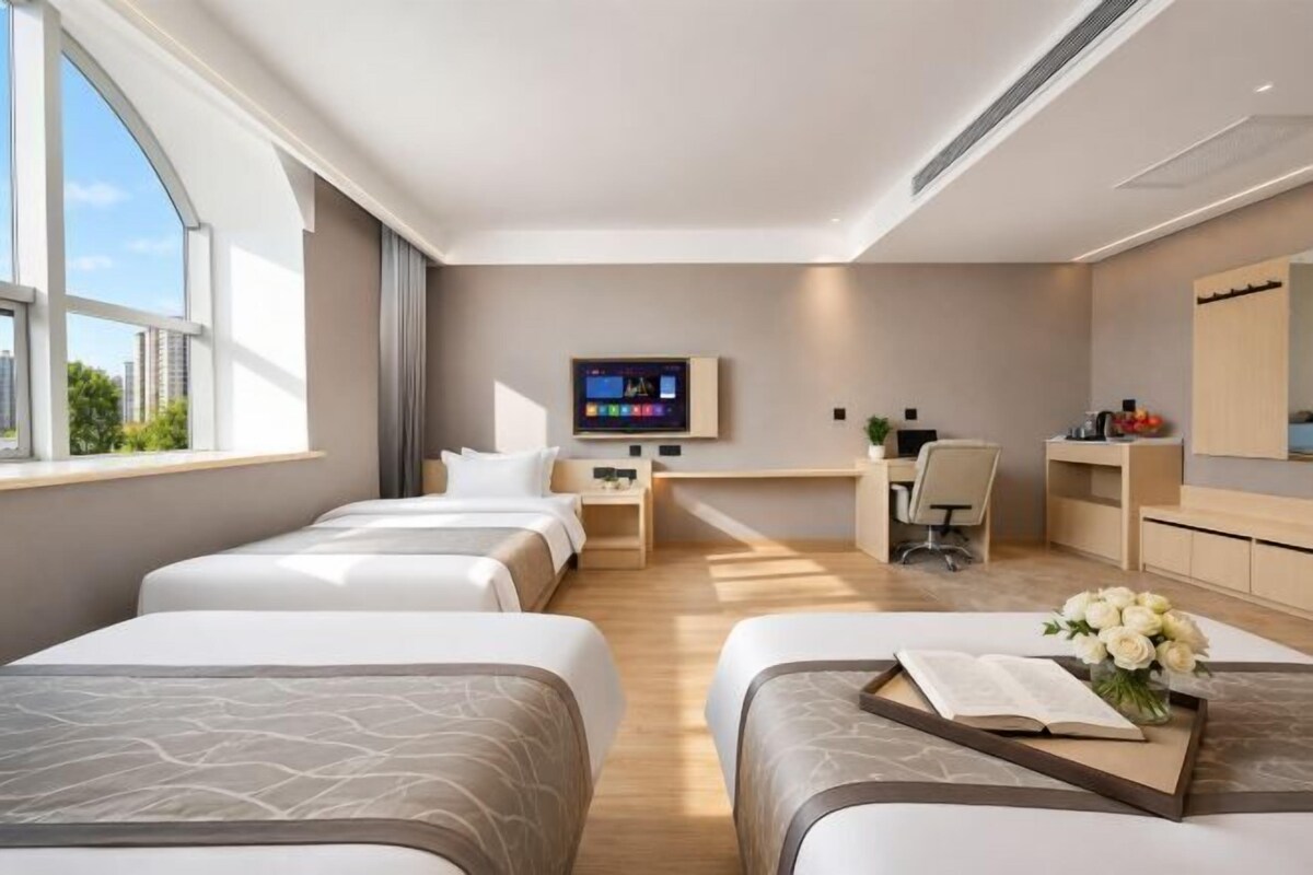 Habitación de hotel con tres camas, televisión, escritorio con silla y una ventana grande.