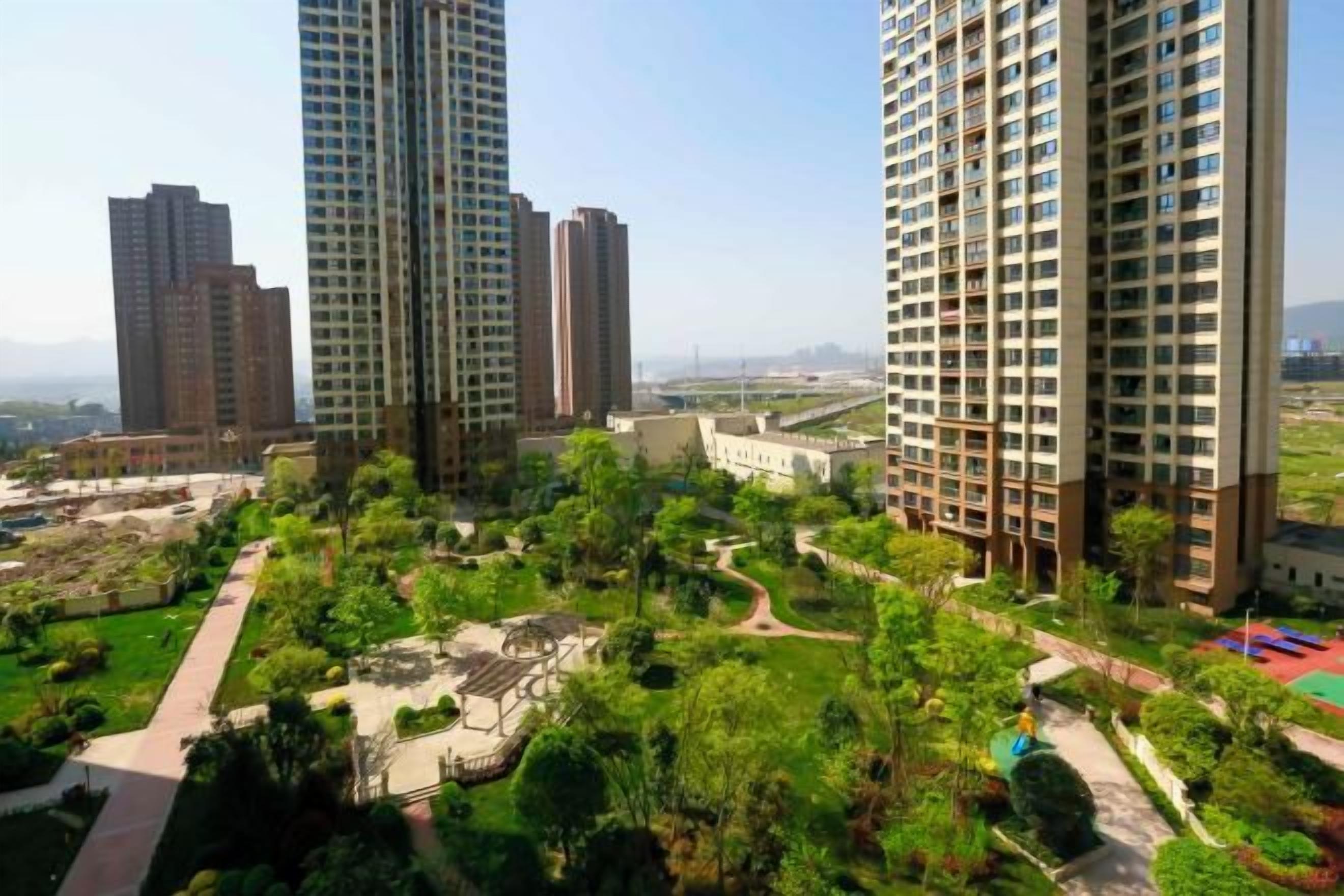 A high-rise residential building complex with multiple apartment buildings, a landscaped garden area, and a clear sky.