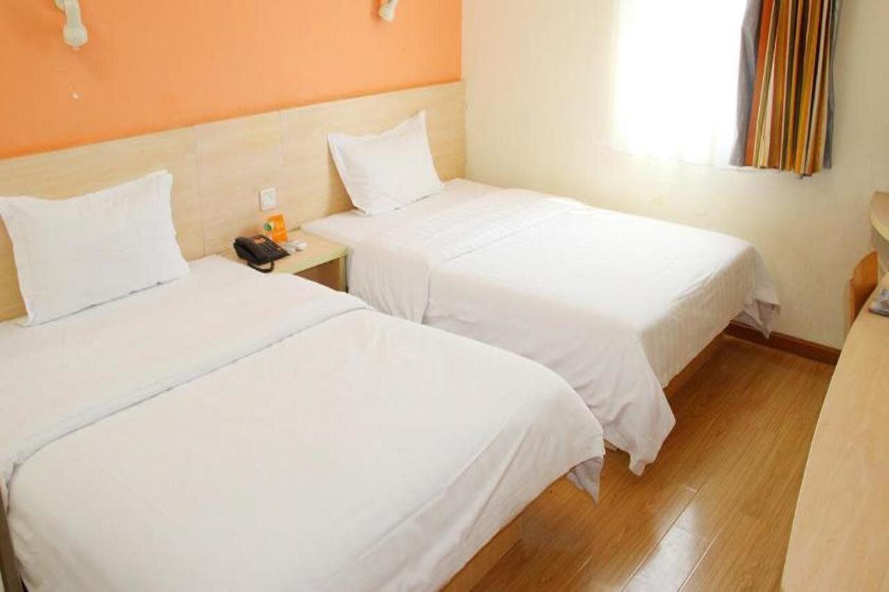 A hotel room with two single beds, a wooden floor, a small bedside table with a phone and a bottle, and a window with curtains.