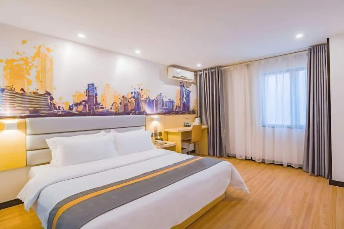 A hotel room with a bed, a desk, a chair, a large window with curtains, and a cityscape painting on the wall.
