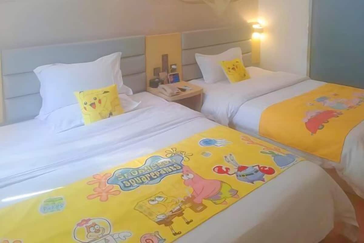 A hotel room with two beds, each with a yellow blanket featuring cartoon characters. There are white pillows and a nightstand with a phone and a lamp.