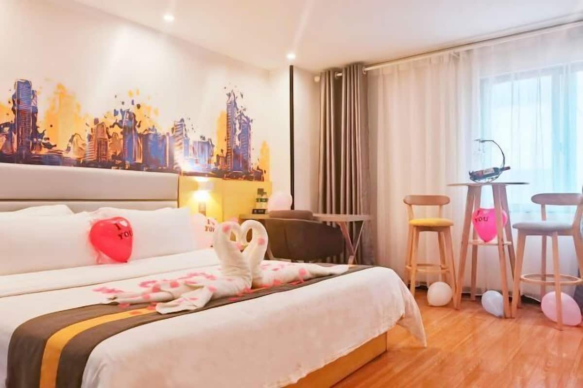 A hotel room with a bed, a cityscape painting, a desk, two chairs, and a window with curtains.