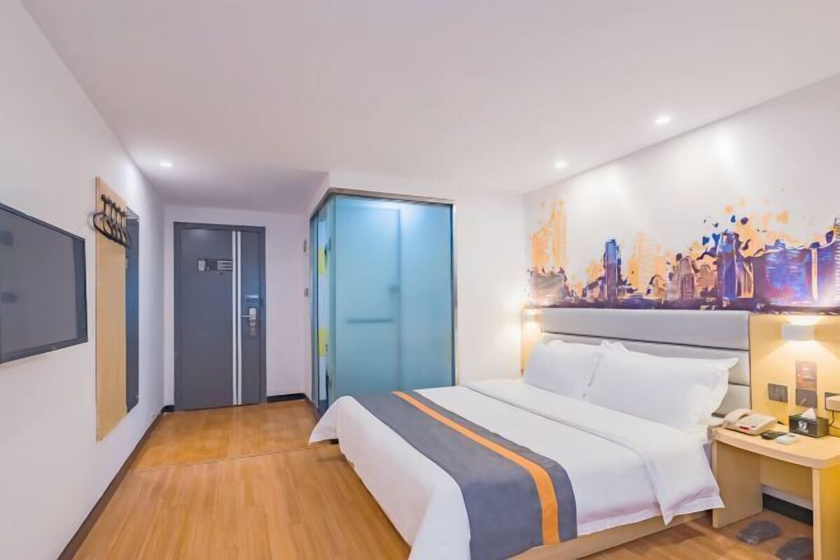 Una habitaci ón de hotel con cama, mesita de noche, televisor montado en la pared, una puerta y un cuadro.