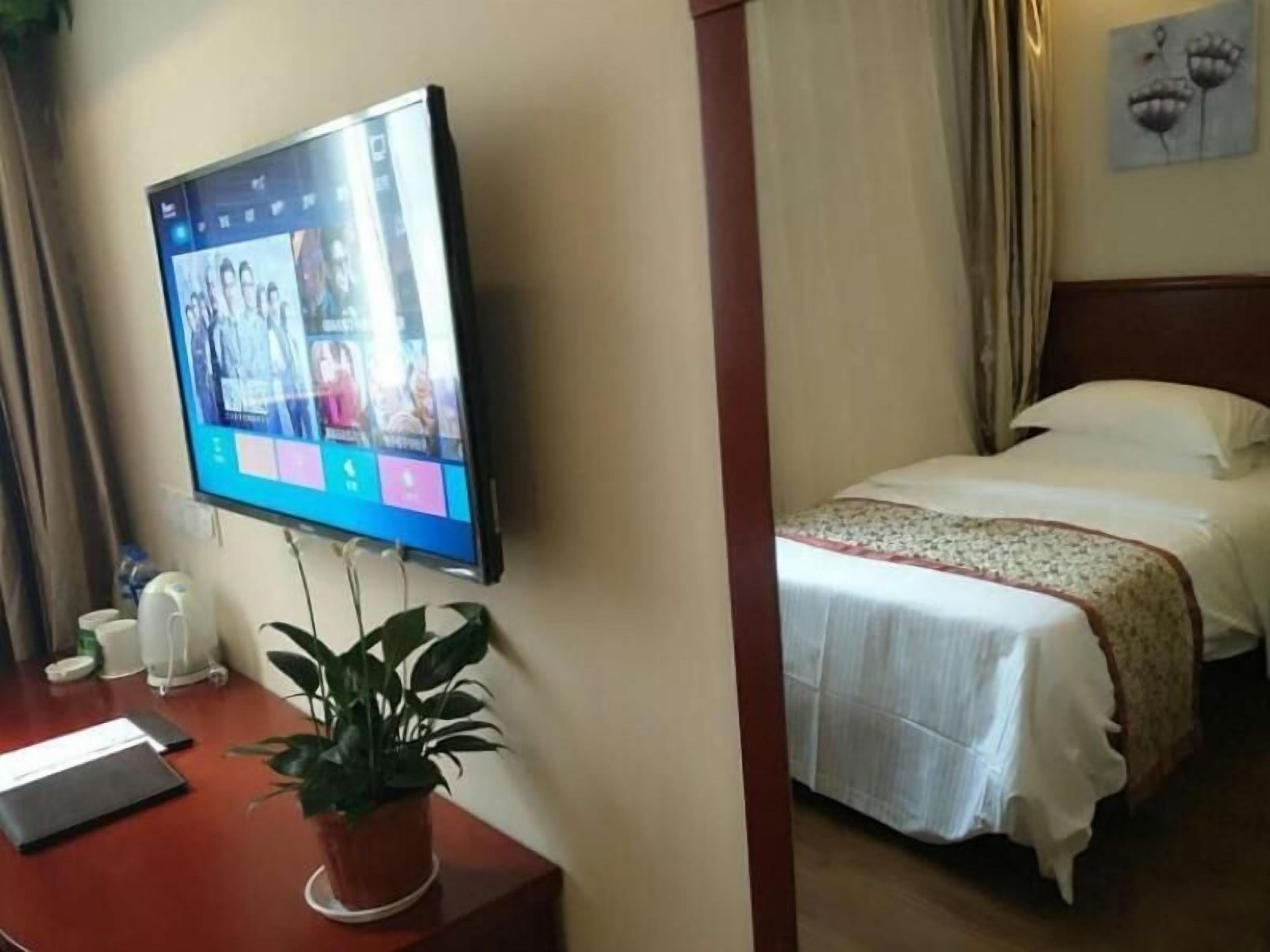 A hotel room with a bed, a desk with a laptop, a potted plant, and a TV mounted on the wall displaying a webpage.