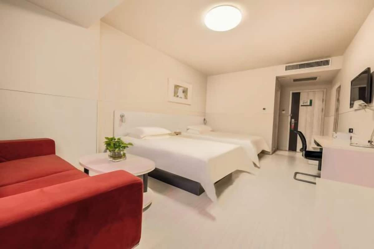 A hotel room with a bed, a red sofa, a small table with a plant, a white bedside table, and a bathroom with a mirror and sink.